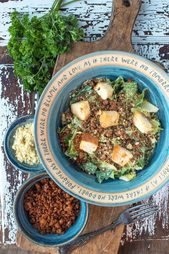 caesar salad in bowl next to vegan bacon bits and vegan parmesan cheese in dishes