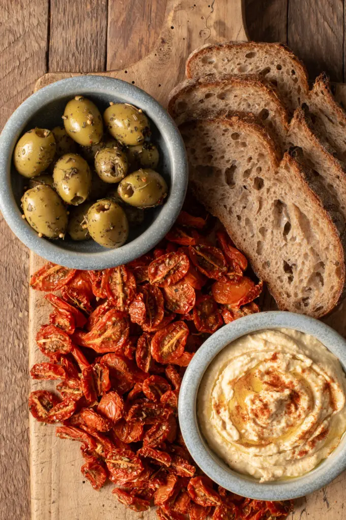 slow roasted cherry tomatoes with hummus, olives and sourdough slices on cutting board