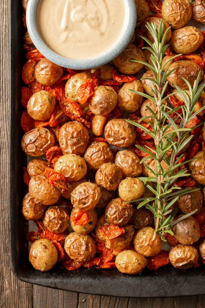roasted potato and tomato tray bake with rosemary sprigs and tahini mustard sauce in dish