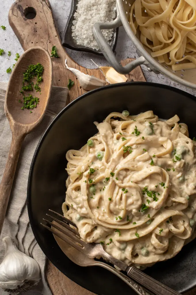 vegan pasta alfredo in black bowl on cutting board next to pasta in strainer