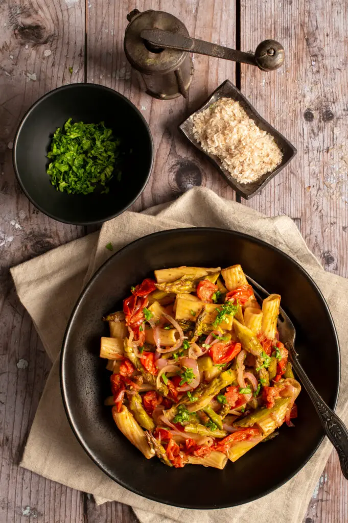 simple asparagus rigatoni in black bowl next to bowls of chopped basil and flaked salt