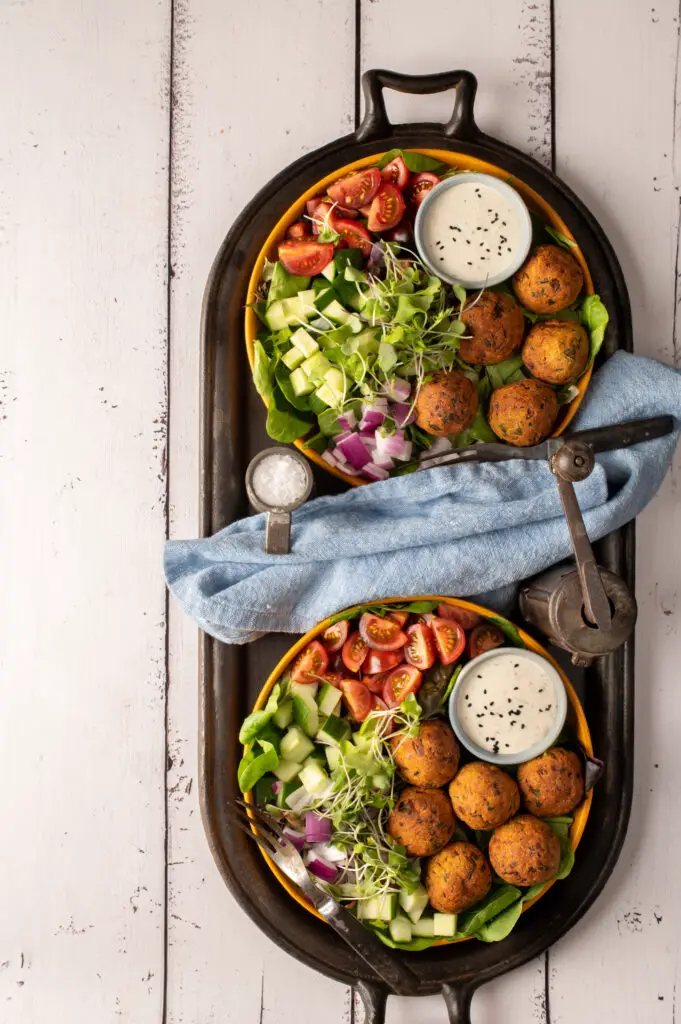 two falafel plates with tomatoes, cucumber and greens sitting on iron griddle